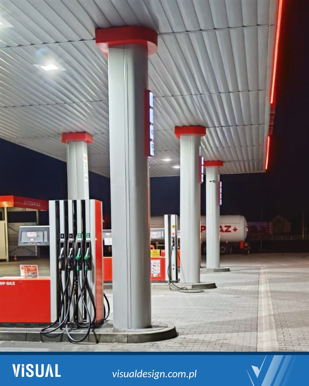 Nowoczesna stacja paliw nocą: białe kolumny z czerwonymi akcentami podświetlone ledami, dystrybutory paliwa i cysterna w tle. Minimalistyczny design.