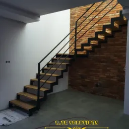 Nowoczesne schody z drewnianymi stopniami i czarną metalową konstrukcją, zamontowane przy ceglanej ścianie, widoczne z dołu, z oświetleniem wpadającym z góry.