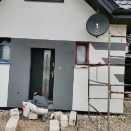 Pracownik siedzi na bloczku betonowym przed budynkiem z dwukolorową elewacją, przygotowując się do prac wykończeniowych przy drzwiach wejściowych. Okna zabezpieczone folią. Widoczne rusztowanie...