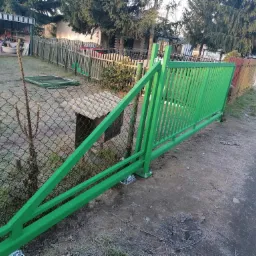 Świeżo pomalowana na zielono przesuwna brama wjazdowa w trakcie montażu, widoczne śruby mocujące do podłoża, w tle ogrodzenie z siatki i drewniany płot.