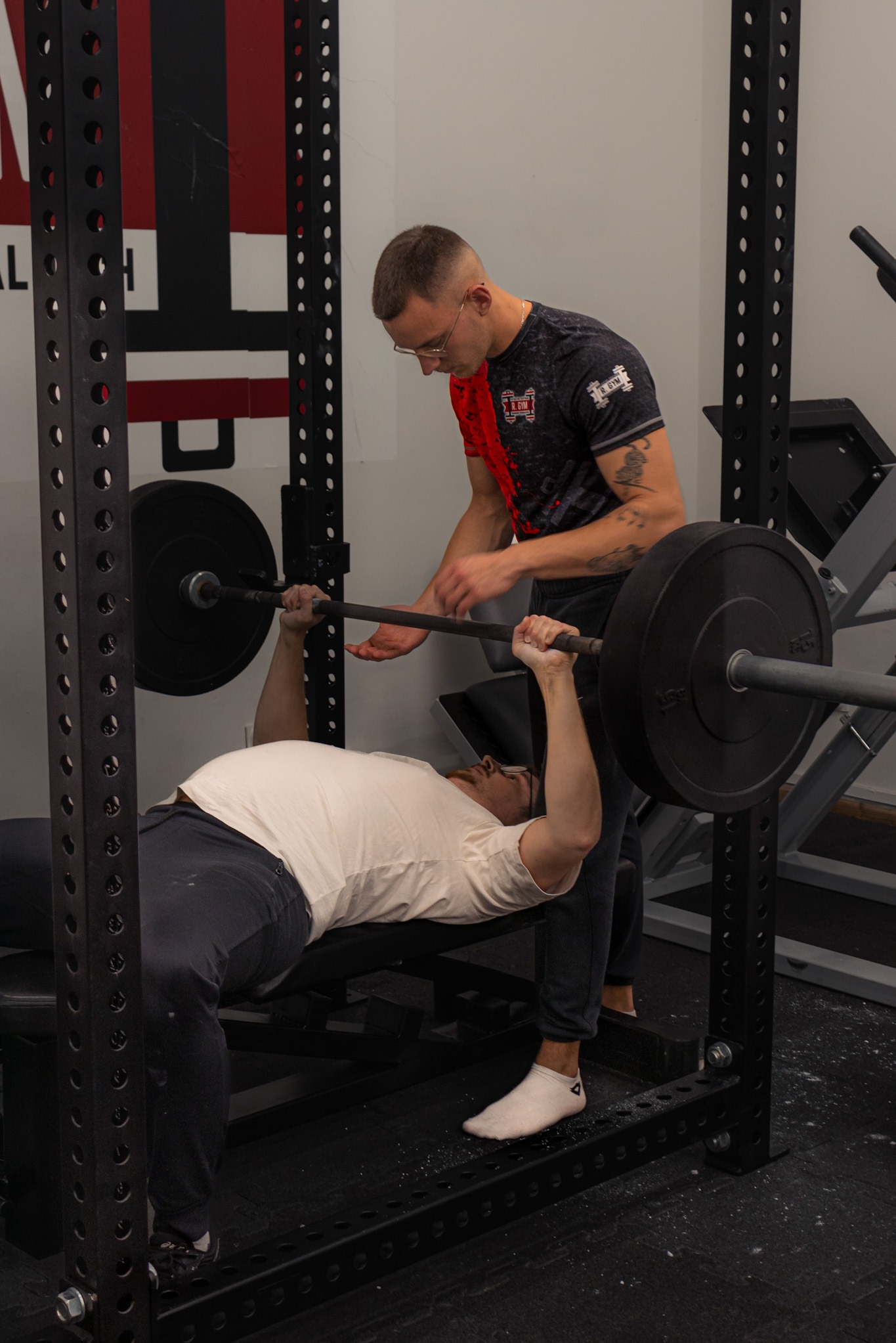 Mężczyzna wyciska sztangę na ławeczce w asyście drugiego mężczyzny w siłowni. Asystent w okularach, z tatuażem na ramieniu, ubrany w koszulkę z logo R.GYM, kontroluje ruch sztangi.