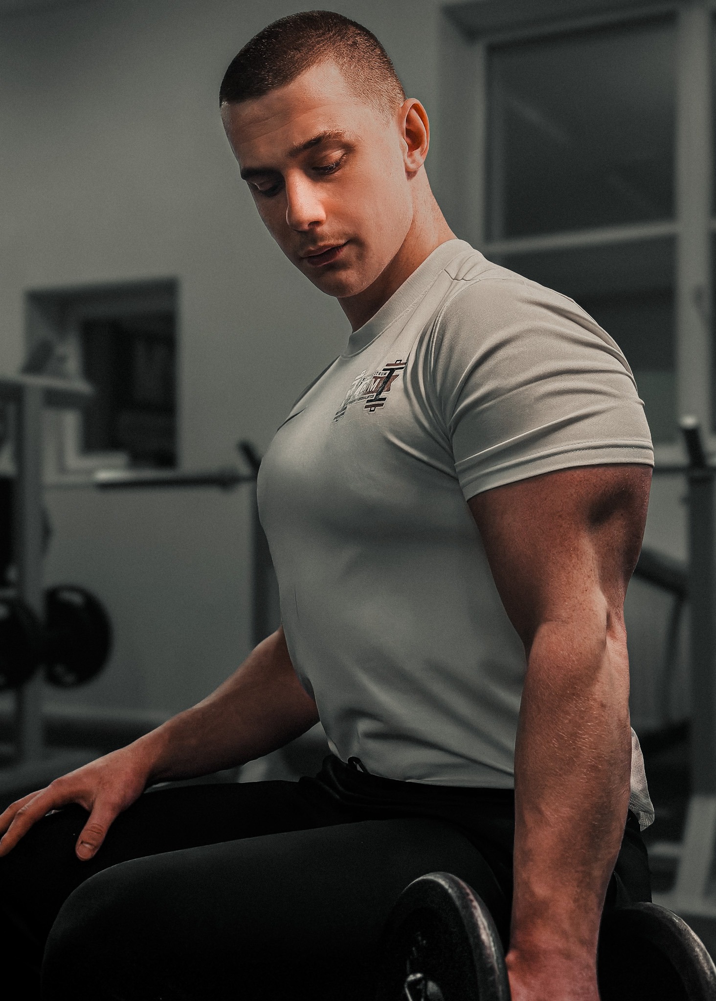Mężczyzna w szarej koszulce siedzi w siłowni, trzymając hantle, z widocznym logo na koszulce i sprzętem do ćwiczeń w tle.