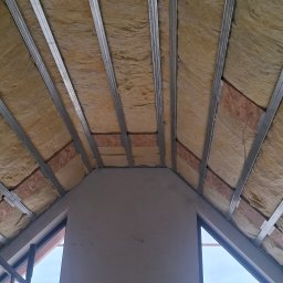 Arkadiusz Wroński Instalacje i Remonty - Skośny sufit poddasza z widoczną izolacją z wełny mineralnej, metalowym stelażem i fragmentem ściany z oknem, przygotowany do montażu płyt.