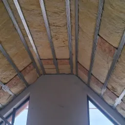 Skośny sufit poddasza z widoczną izolacją z wełny mineralnej, metalowym stelażem i fragmentem ściany z oknem, przygotowany do montażu płyt.