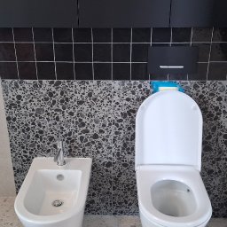 Arkadiusz Wroński Instalacje i Remonty - Nowoczesna łazienka z białą armaturą, czarnymi płytkami i mozaiką. Widoczny bidet, toaleta i fragment kabiny prysznicowej. Minimalistyczny design z dbałością o detale.