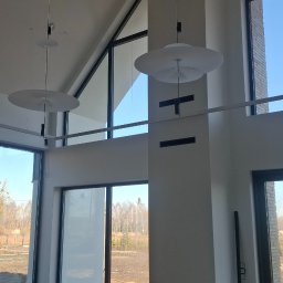 Arkadiusz Wroński Instalacje i Remonty - Wysokie wnętrze z nowoczesnym oświetleniem: dwa okrągłe plafony i szyna z reflektorami. Widoczna drabina, duże okna z widokiem na zewnątrz. Minimalistyczny design.