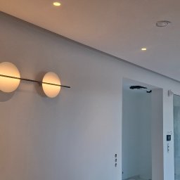Arkadiusz Wroński Instalacje i Remonty - Nowoczesne oświetlenie ścienne: trzy okrągłe lampy z poziomą listwą w minimalistycznym wnętrzu. Widoczne także punktowe światła sufitowe i fragment korytarza.