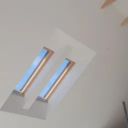 Wnętrze poddasza z dwoma oknami dachowymi skierowanymi w górę, widoczne drewniane elementy konstrukcyjne dachu i fragment drabiny malarskiej.