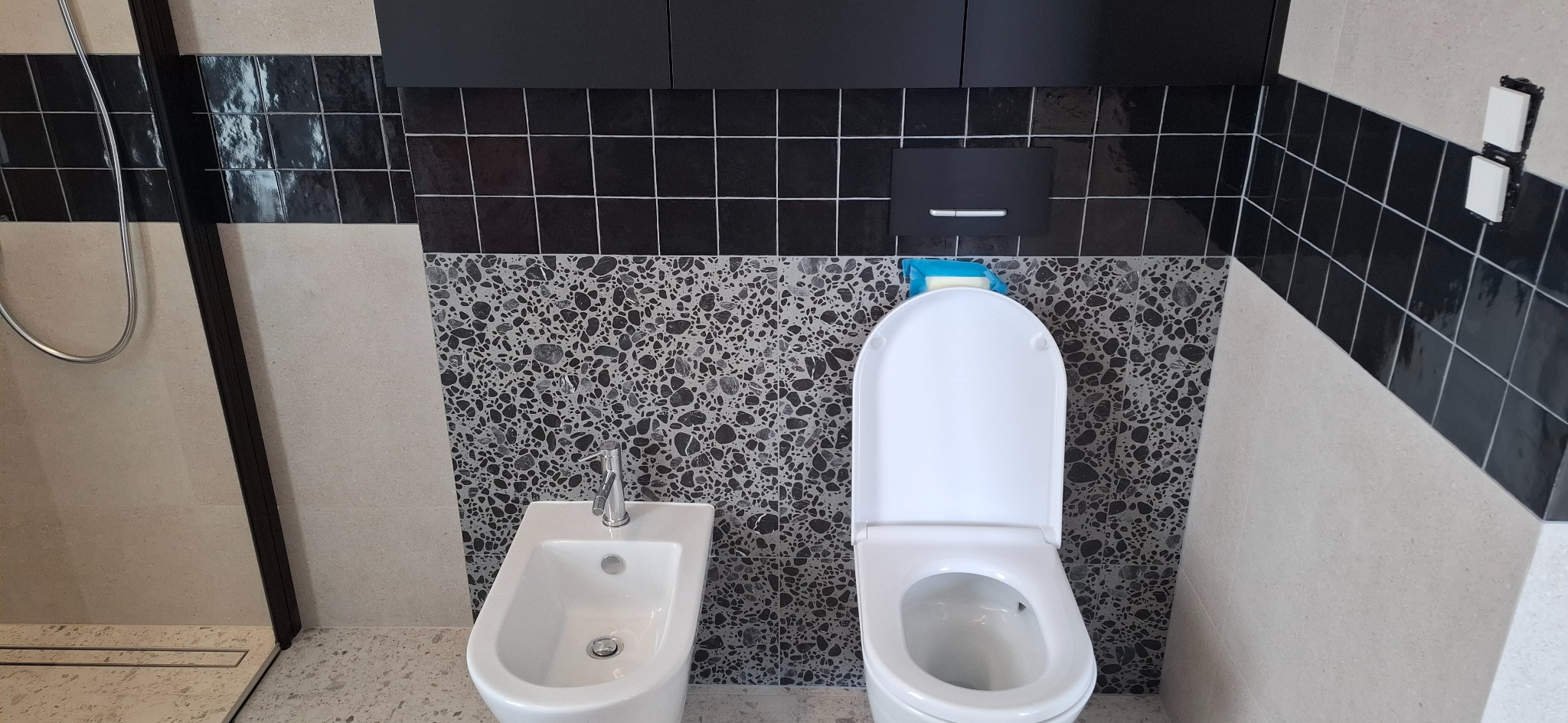 Nowoczesna łazienka z białą armaturą, czarnymi płytkami i mozaiką. Widoczny bidet, toaleta i fragment kabiny prysznicowej. Minimalistyczny design z dbałością o detale.