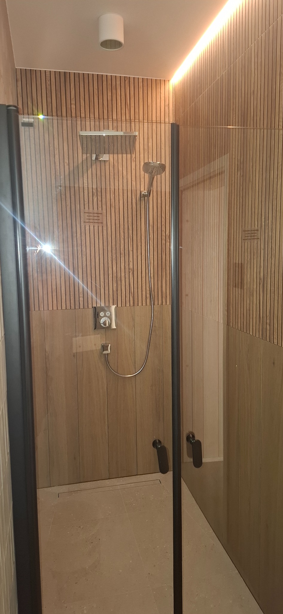 Nowoczesna kabina prysznicowa z deszczownicą i panelami ściennymi imitującymi drewno. Minimalistyczny design, stonowana kolorystyka, oświetlenie LED.
