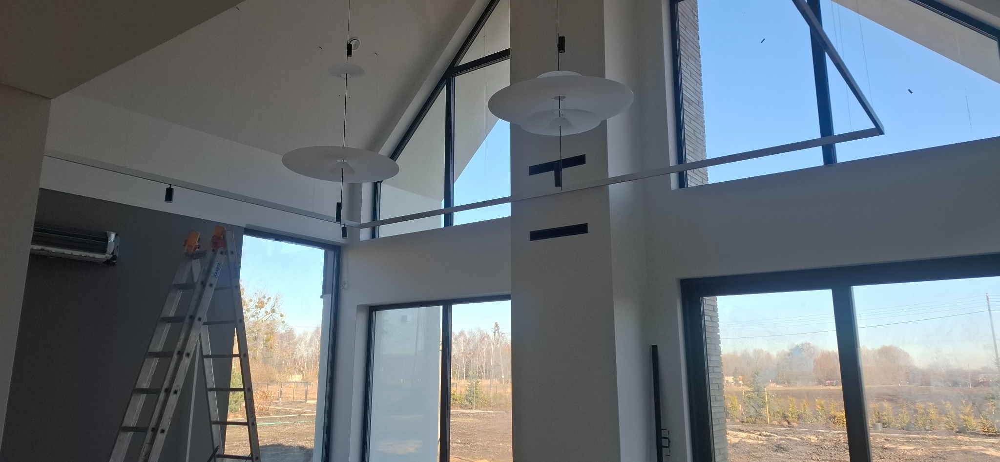 Wysokie wnętrze z nowoczesnym oświetleniem: dwa okrągłe plafony i szyna z reflektorami. Widoczna drabina, duże okna z widokiem na zewnątrz. Minimalistyczny design.