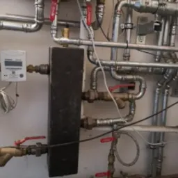 Skomplikowana instalacja rur miedzianych z zaworami i licznikiem ciepła na ścianie w kotłowni, widoczne złącza lutowane i skręcane.