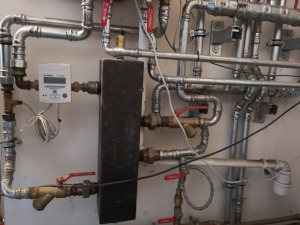 Skomplikowana instalacja rur miedzianych z zaworami i licznikiem ciepła na ścianie w kotłowni, widoczne złącza lutowane i skręcane.