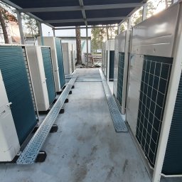 Klimacraft Robert Kusz, Rafał Pospiech - Rząd zewnętrznych jednostek klimatyzacyjnych, ustawionych pod zadaszeniem na betonowym podłożu, z widocznymi korytkami kablowymi i budynkiem w tle.