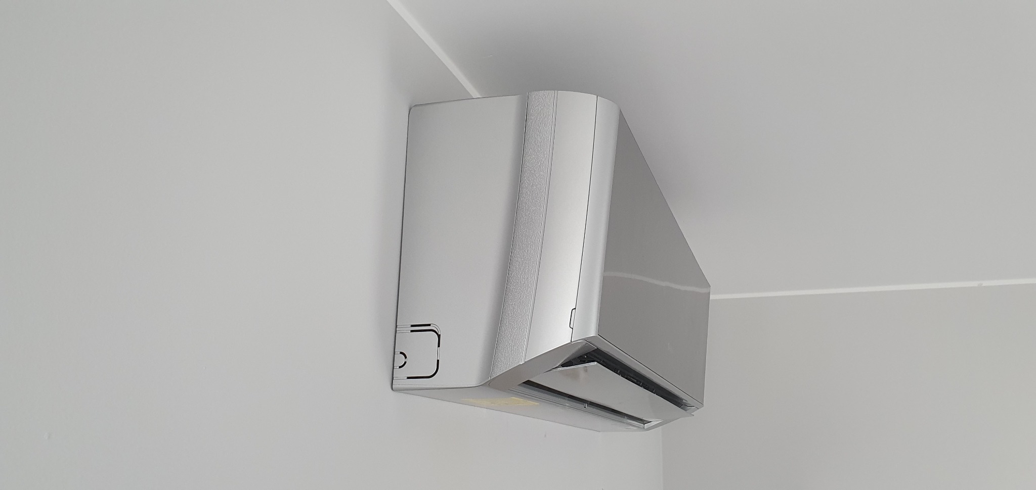 Nowoczesny, srebrny klimatyzator ścienny zamontowany w rogu pomieszczenia o białych ścianach i suficie. Widoczny fragment listwy sufitowej.
