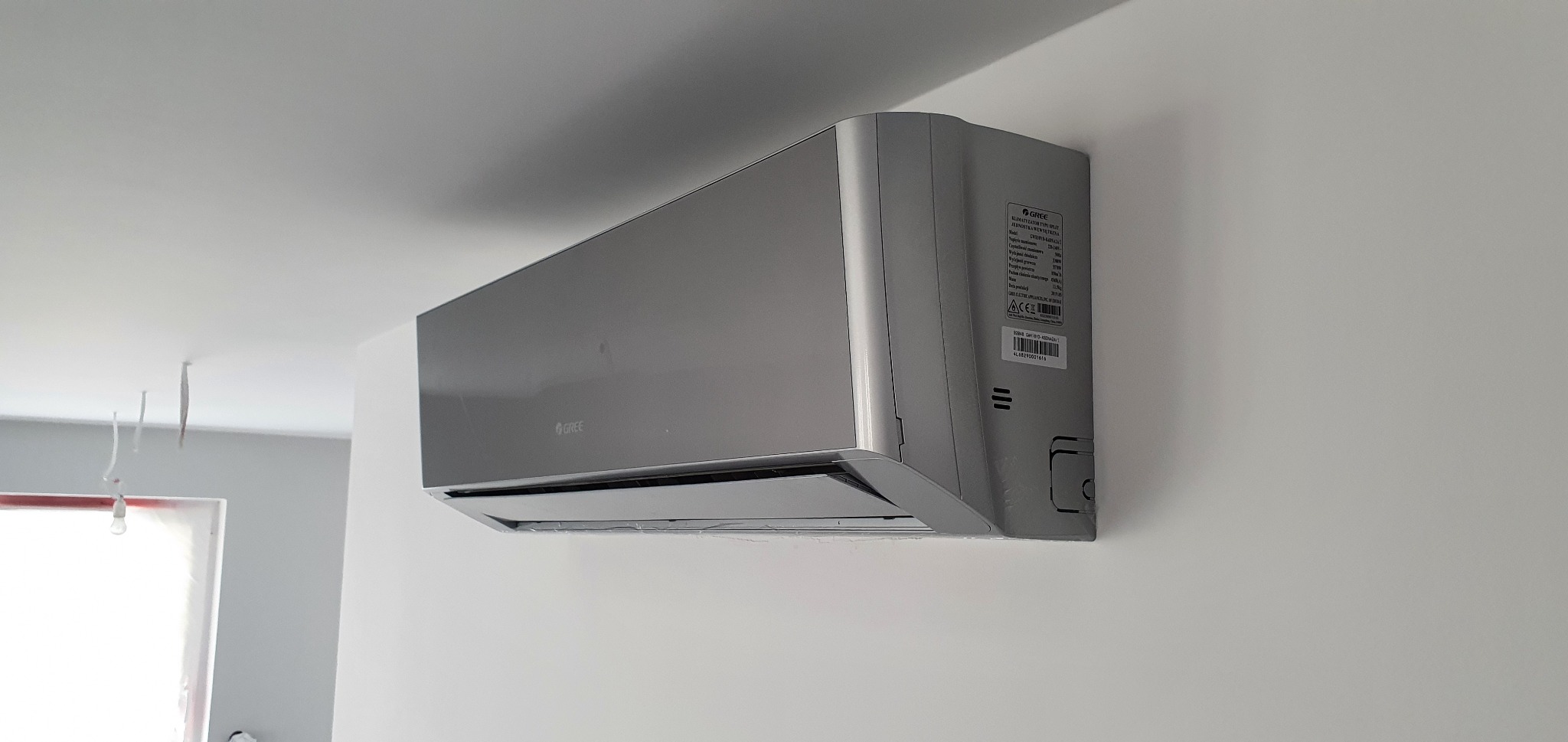 Szary klimatyzator Gree zamontowany na białej ścianie w pomieszczeniu w trakcie remontu, widoczne wiszące kable i zakryte okno.