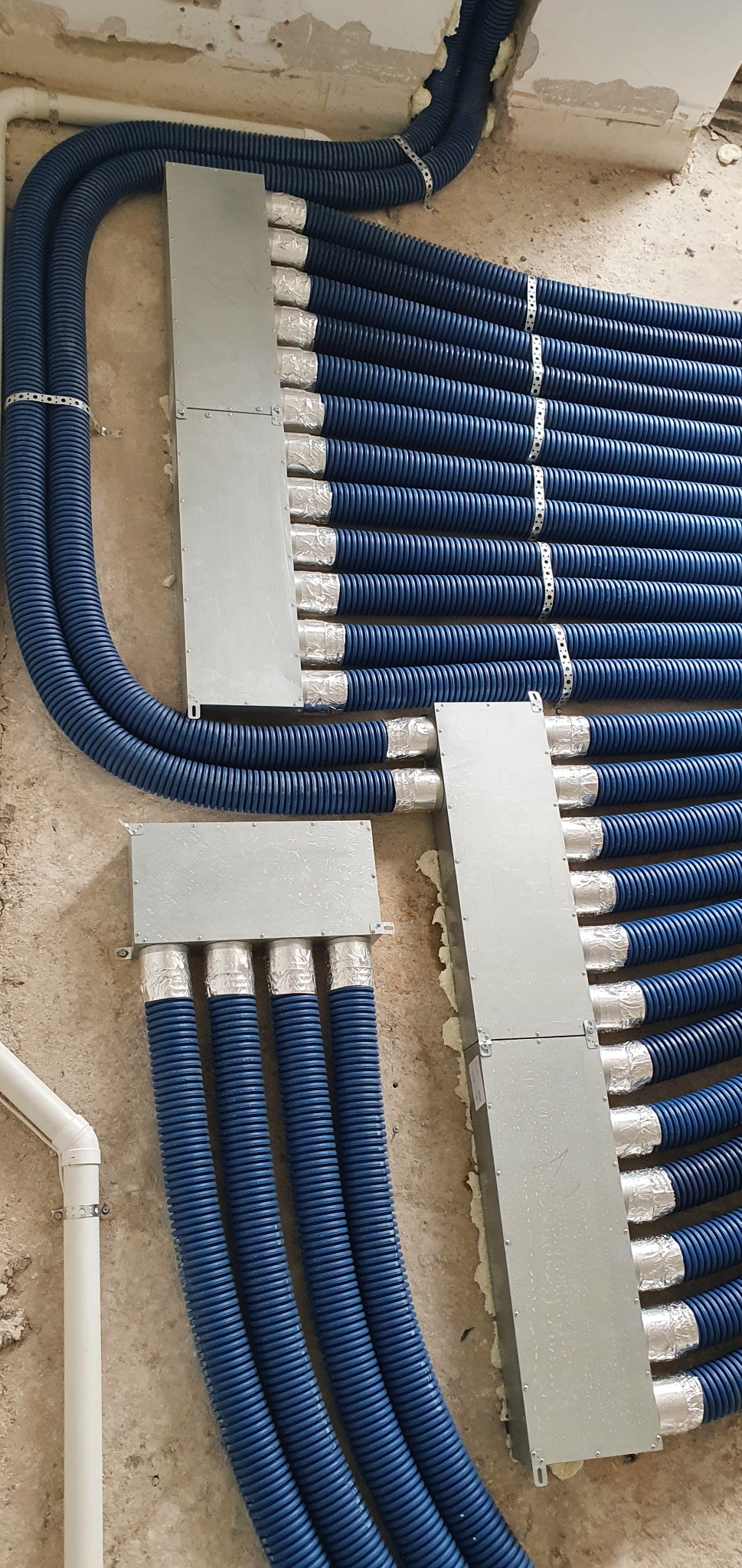 Równolegle ułożone, niebieskie, karbowane rury wentylacyjne przymocowane do surowej ściany za pomocą metalowych obejm i puszek, z izolacją z folii aluminiowej na końcach.