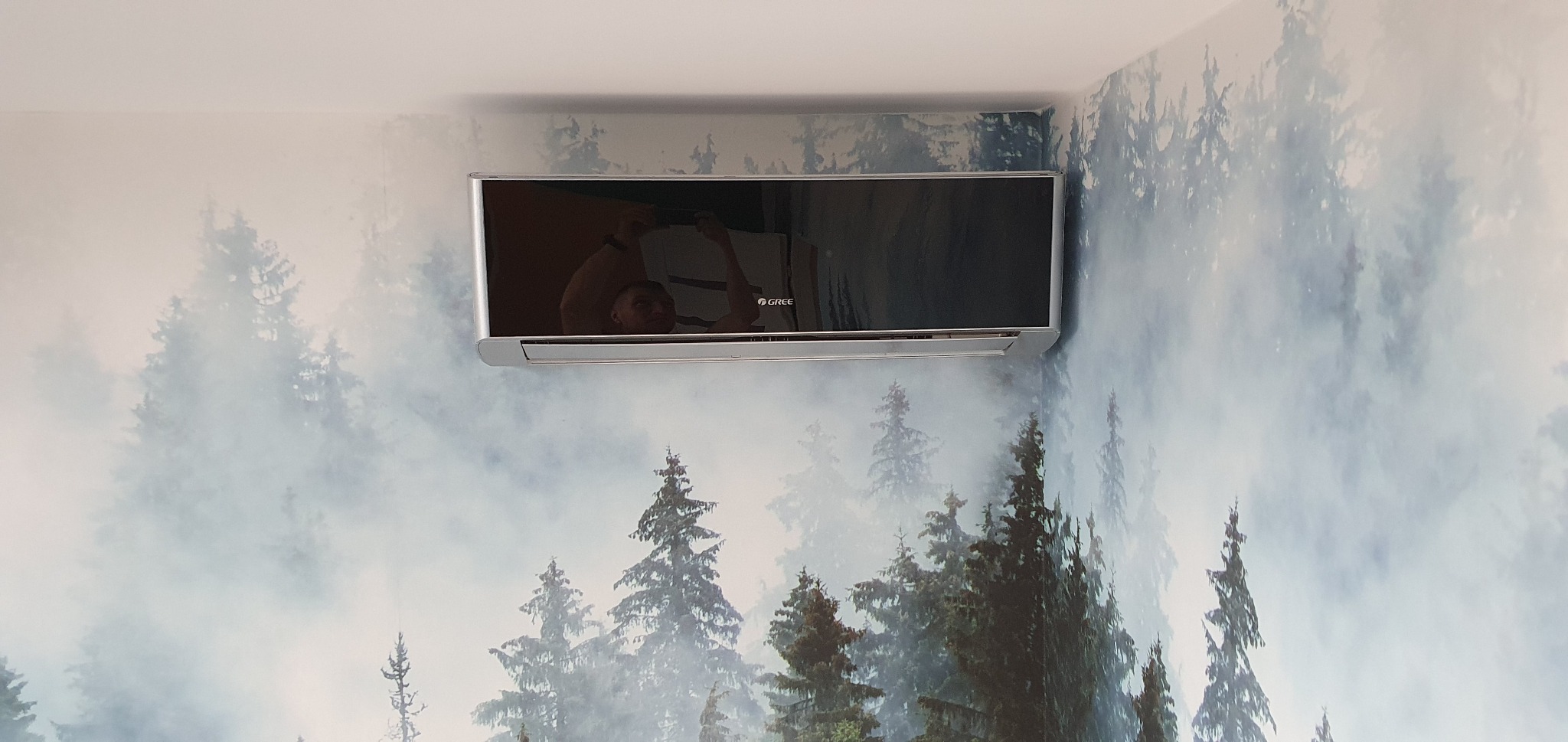 Zamontowany klimatyzator marki Gree na ścianie z fototapetą przedstawiającą las we mgle, widoczny w Suchym Lesie, z odbiciem osoby robiącej zdjęcie w czarnej obudowie klimatyzatora.