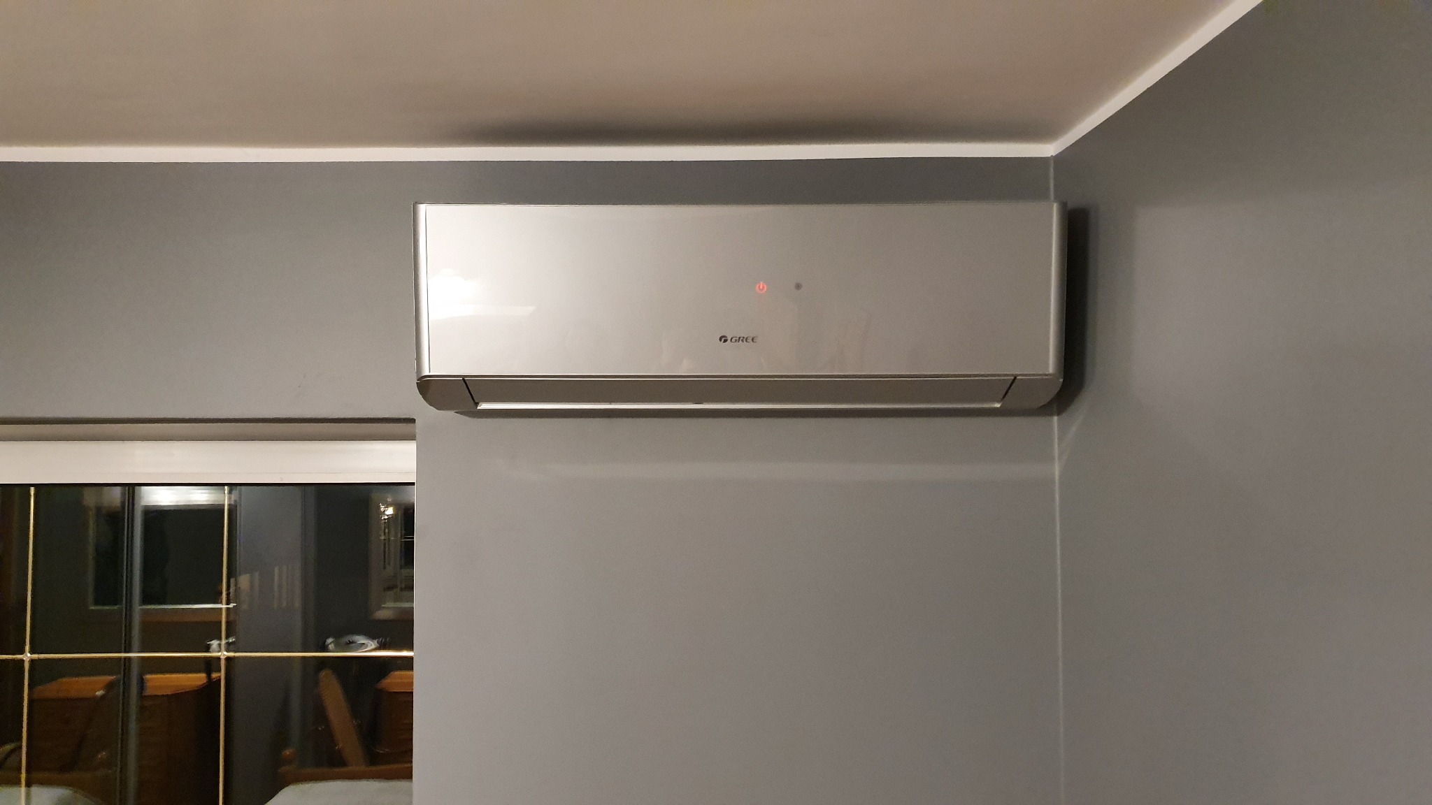 Klimatyzator marki Gree zamontowany na szarej ścianie w pokoju, widoczny przez okno fragment sypialni.