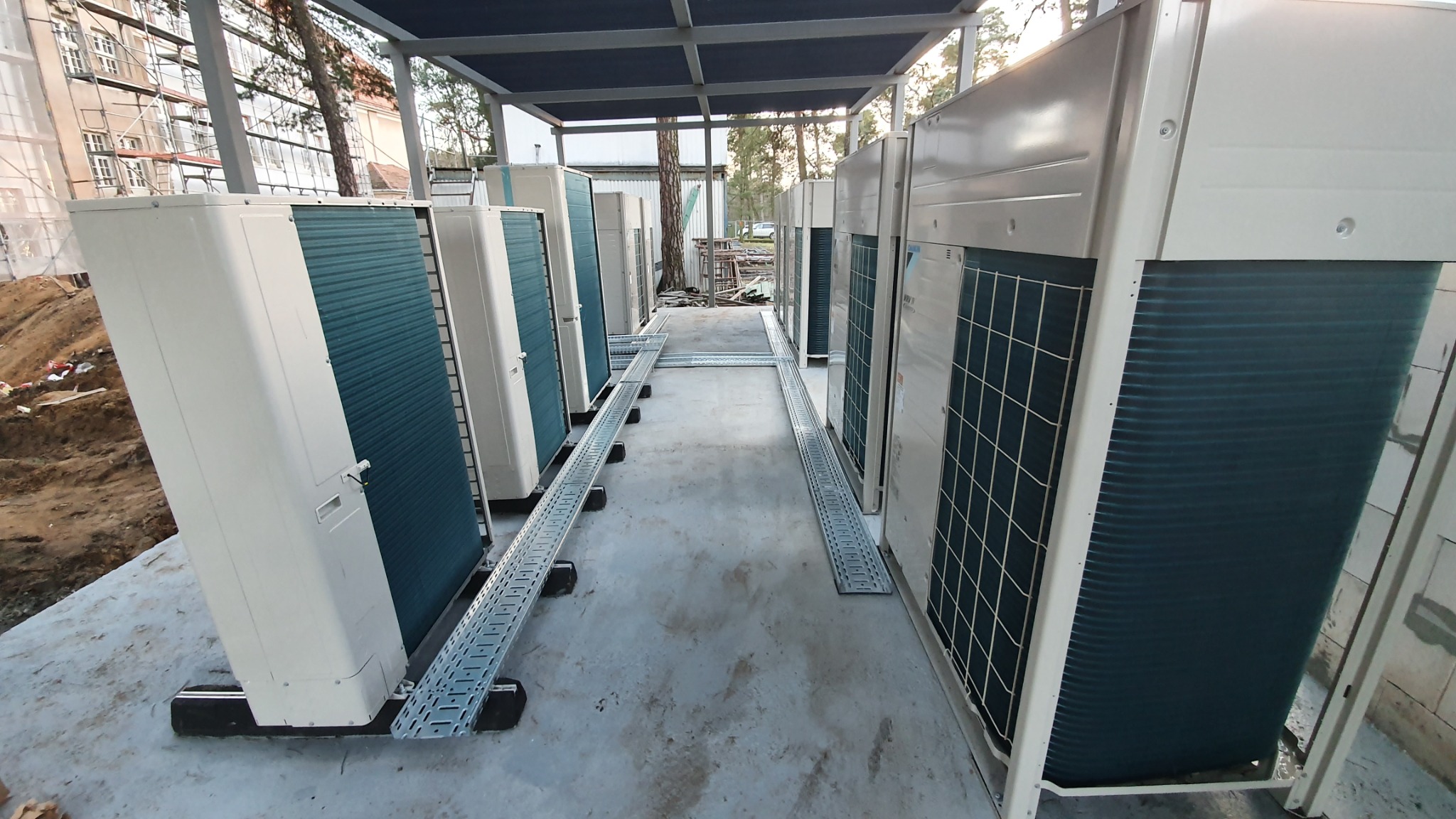 Rząd zewnętrznych jednostek klimatyzacyjnych, ustawionych pod zadaszeniem na betonowym podłożu, z widocznymi korytkami kablowymi i budynkiem w tle.