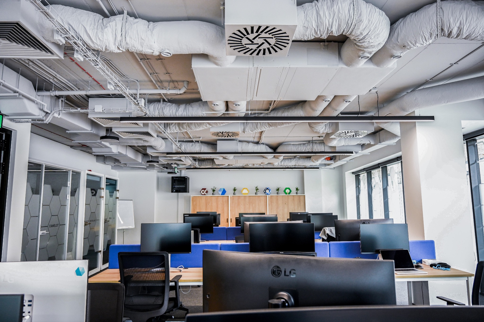 Nowoczesne biuro z widoczną instalacją wentylacyjną pod sufitem, biurkami z monitorami i szklanymi ściankami działowymi. Minimalistyczny design.