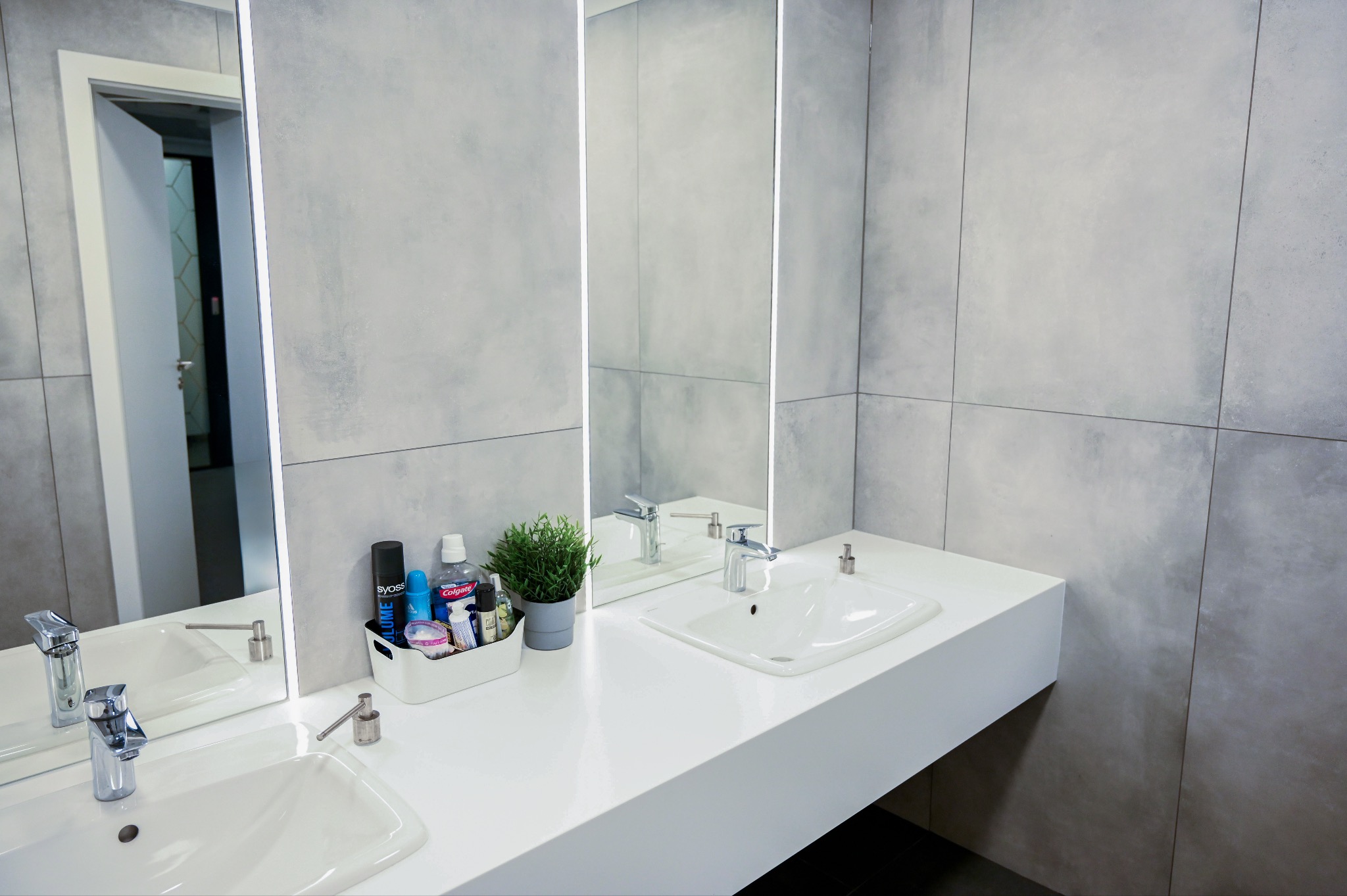 Nowoczesna łazienka biurowa z szarymi płytkami, podwójną białą umywalką i dużym lustrem z podświetleniem LED. Widoczna armatura i akcesoria łazienkowe.