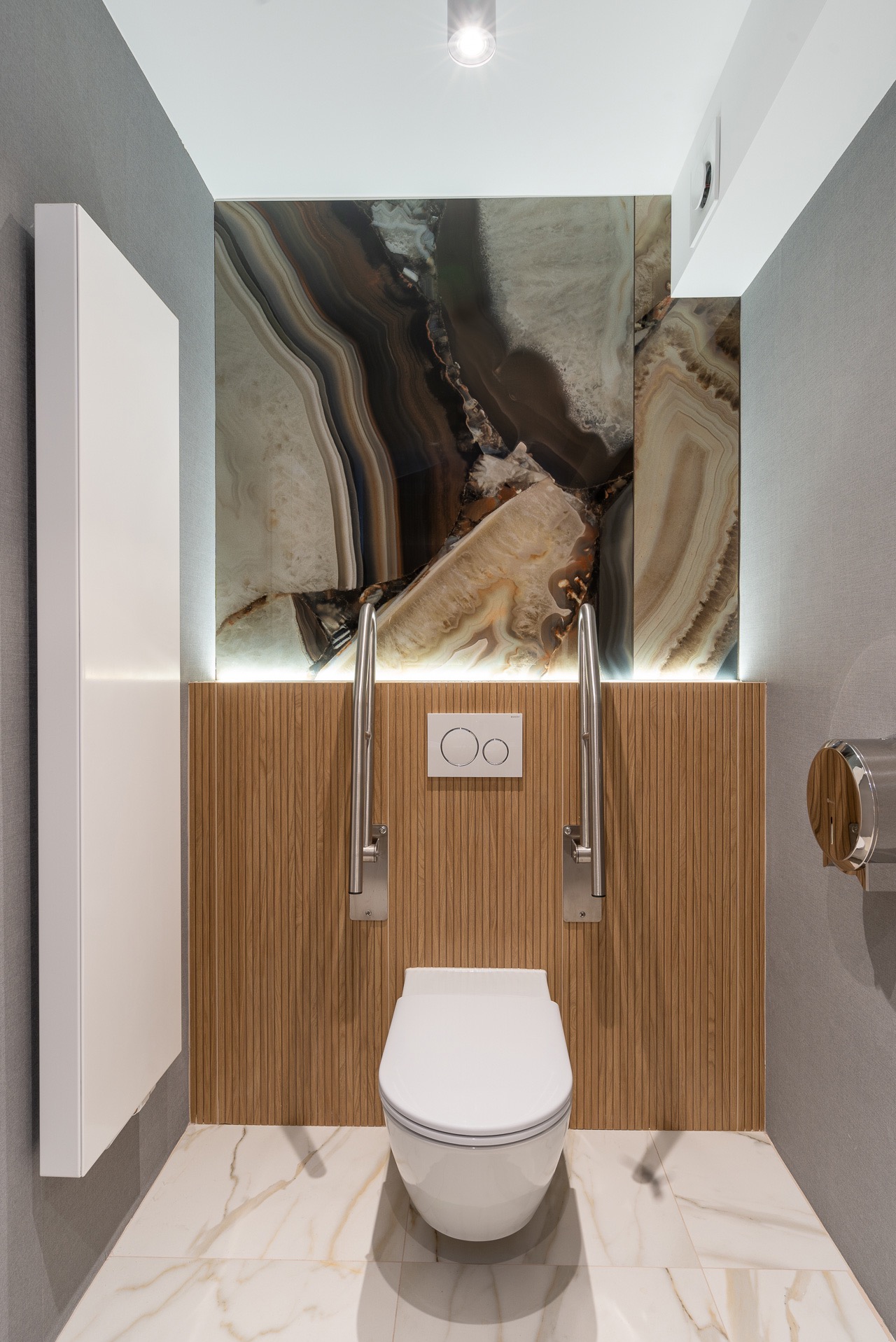 Elegancka toaleta z marmurową ścianą i drewnianą boazerią, wyposażona w uchwyty dla niepełnosprawnych. Oświetlenie LED podkreśla unikalny wzór kamienia.