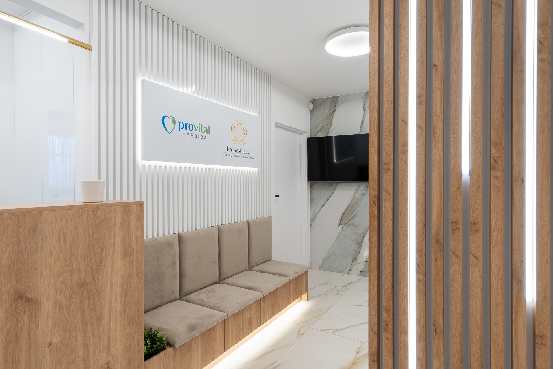 Nowoczesne wnętrze biura z recepcją, sofą i ścianą z logo firmy Provital Medica i ProAesthetic, z elementami drewnianymi i marmurowymi.