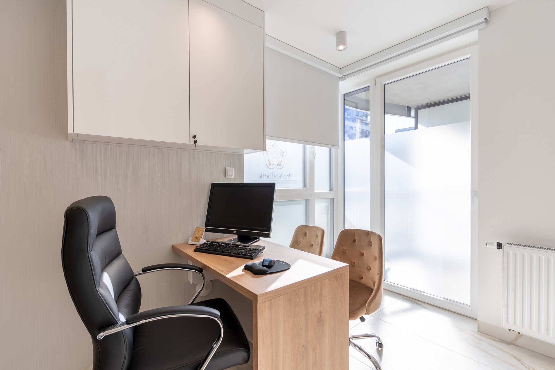 Nowoczesne biuro z drewnianym biurkiem, czarnym krzesłem i dużym oknem z roletami. Na biurku komputer, klawiatura i myszka. Białe szafki wiszące i jasna podłoga.