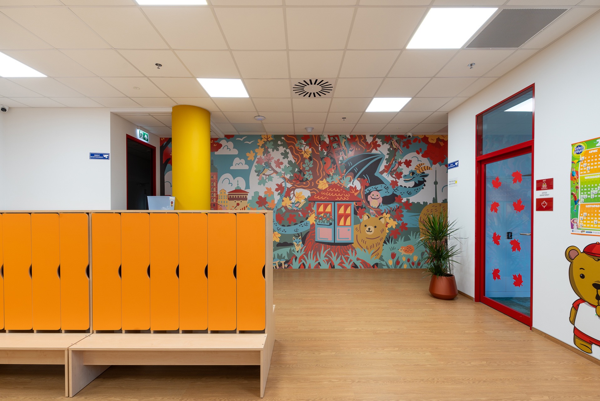 Kolorowe wnętrze biura lub przedszkola: pomarańczowe szafki, bajkowy mural na ścianie, drewniana podłoga i czerwone drzwi. Jasne oświetlenie.