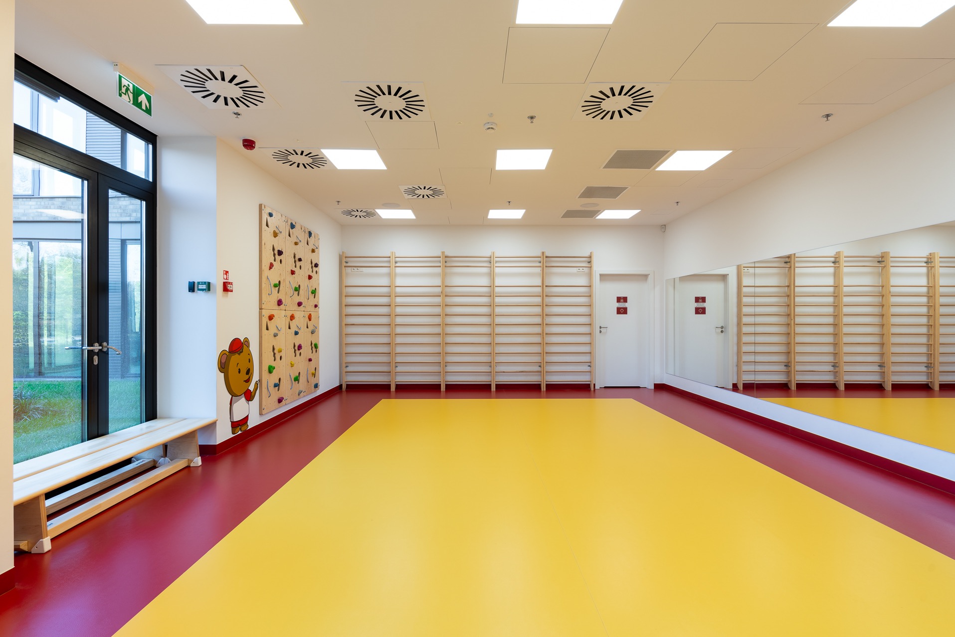 Jasna sala gimnastyczna z drabinkami, ścianką wspinaczkową z misiami i żółto-czerwoną podłogą. Duże okno i lustro na ścianie.