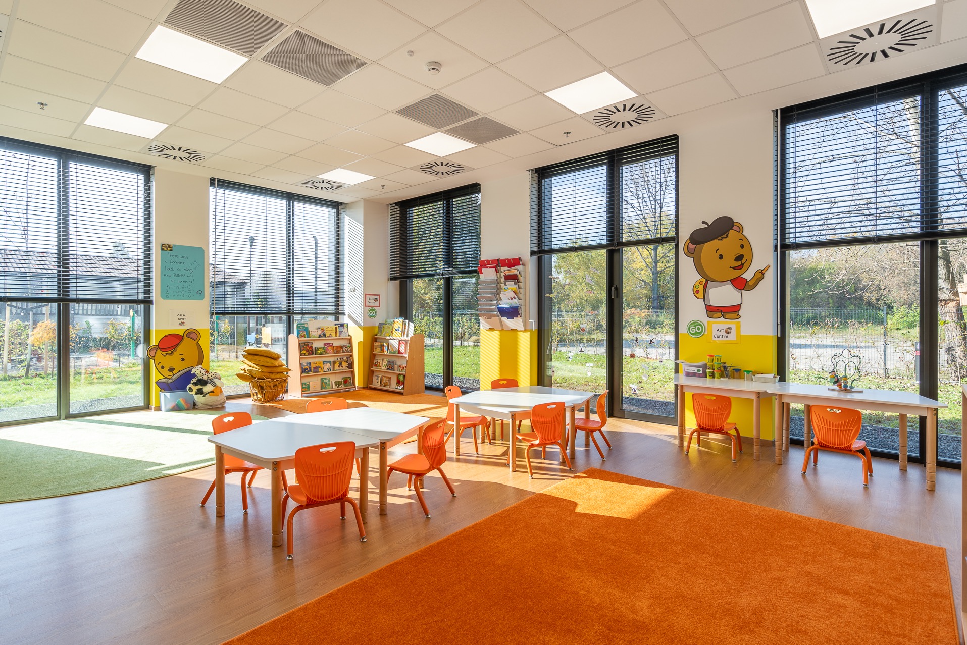 Jasne, nowocześnie urządzone wnętrze przedszkola z pomarańczowymi akcentami: krzesła, dywan. Białe stoliki, regały z książkami i duże okna z widokiem na zewnątrz.