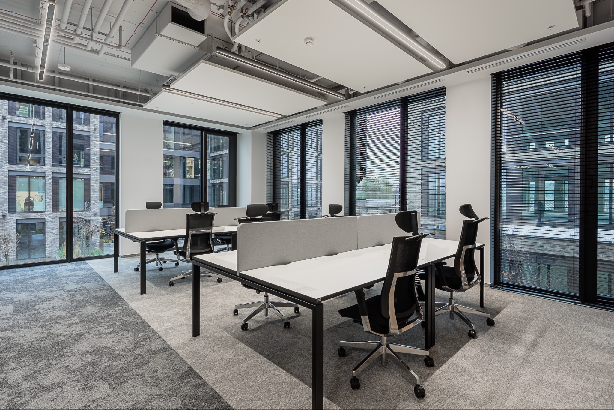 Jasne biuro z biurkami i krzesłami, widok na budynek przez okna z żaluzjami. Nowoczesne oświetlenie sufitowe i wykładzina w neutralnych odcieniach szarości.