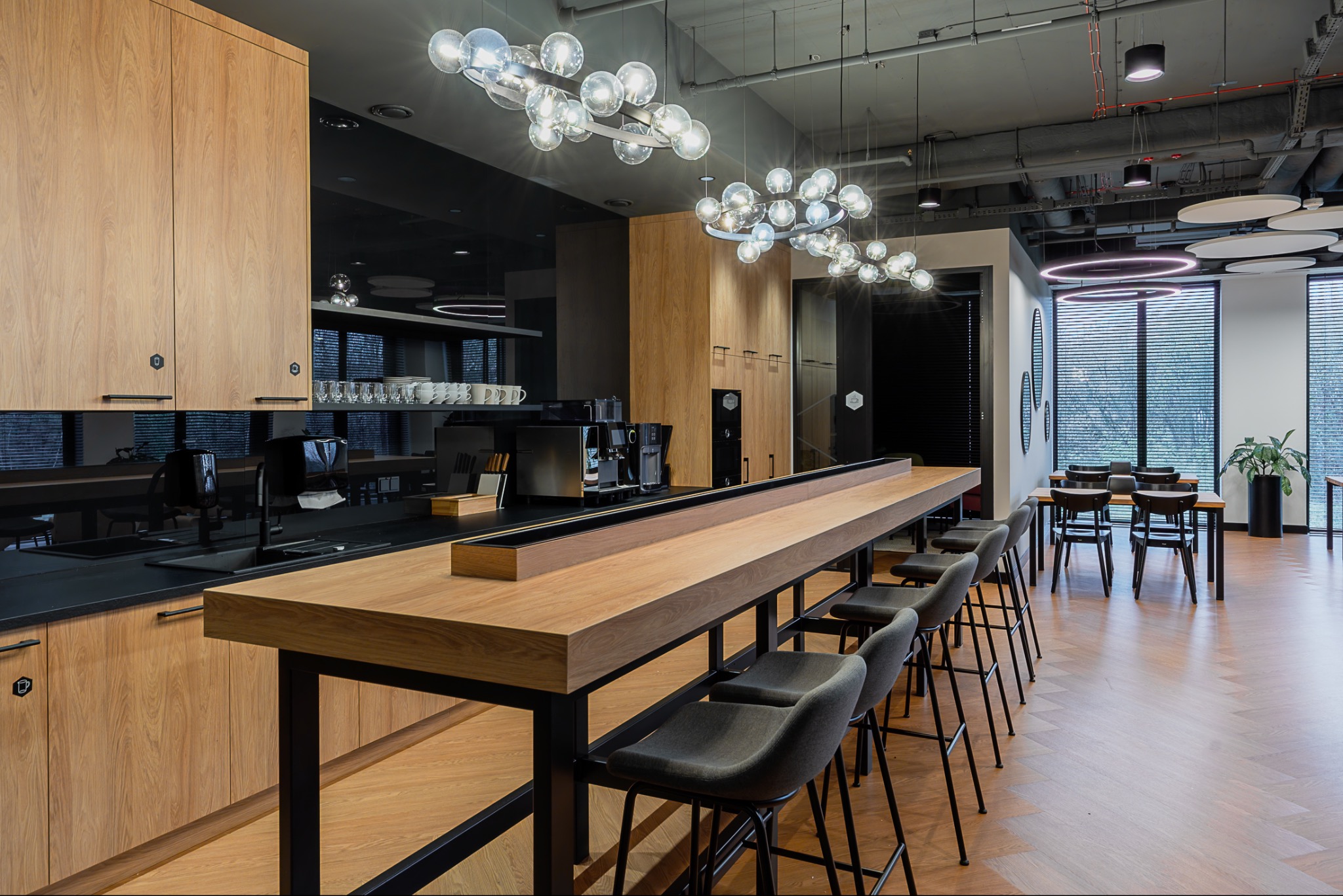 Nowoczesna przestrzeń biurowa z drewnianymi szafkami, czarnym blatem i designerskim oświetleniem. Ustawione krzesła barowe i stoły tworzą strefę relaksu.