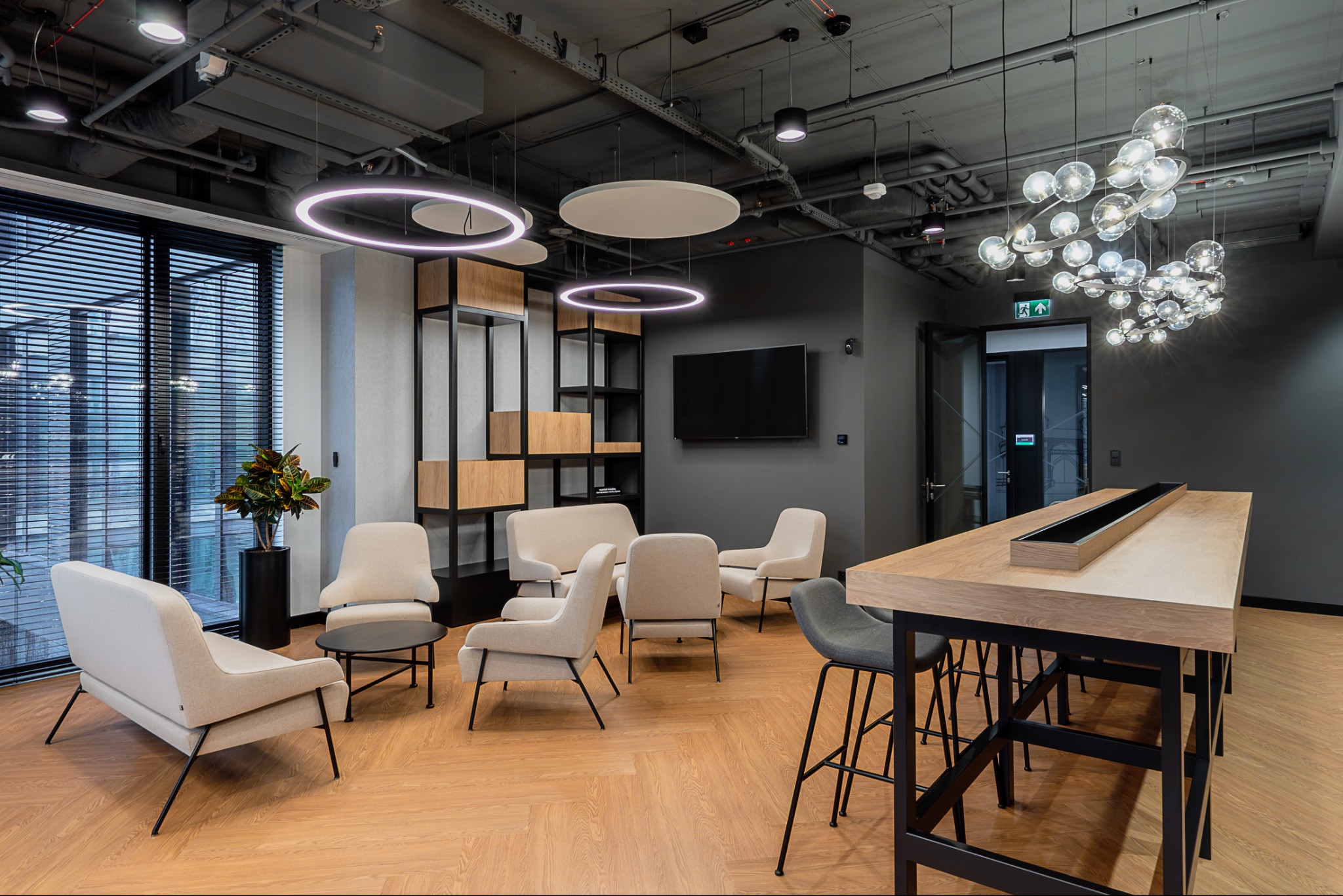 Nowoczesna aranżacja biura: jasne fotele, długa drewniana ława, okrągłe lampy sufitowe i designerski żyrandol w przestronnym wnętrzu.