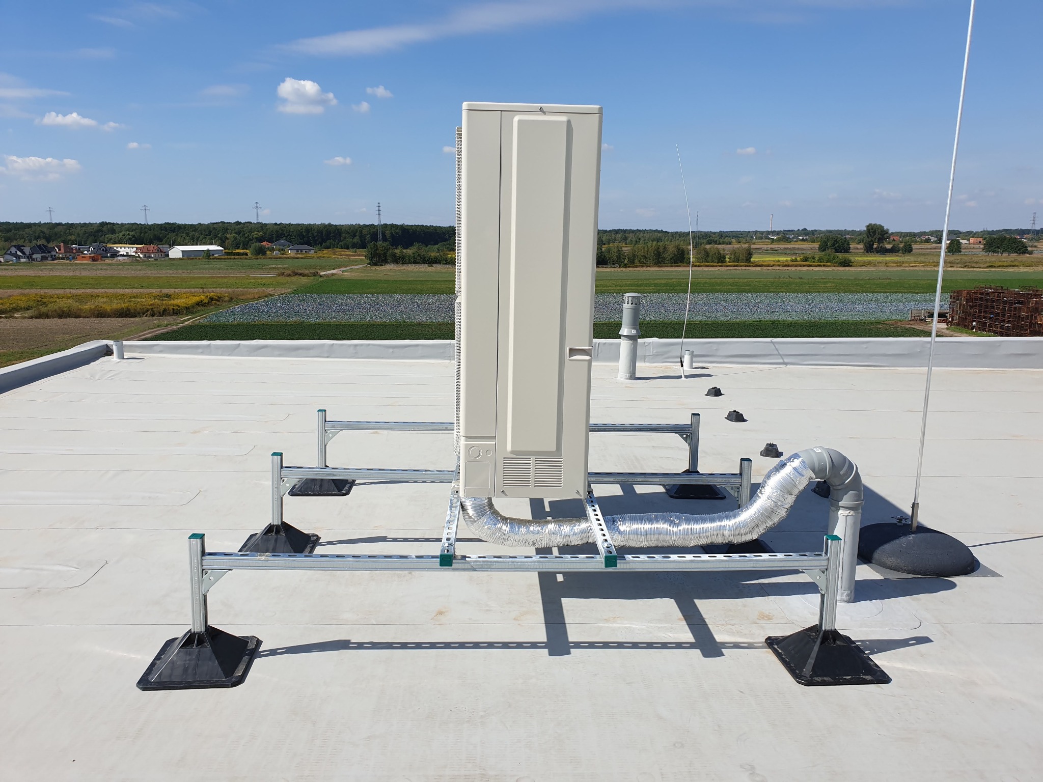 Kompaktowa jednostka klimatyzacyjna na płaskim dachu, zamontowana na metalowej konstrukcji wsporczej, z widocznym elastycznym przewodem wentylacyjnym i anteną, na tle zielonych pól i błękitnego nieba.