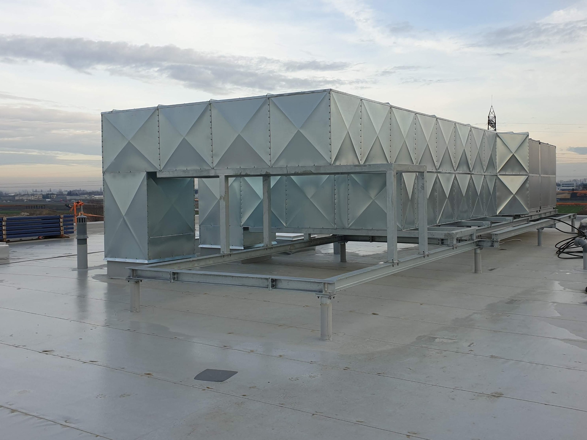 Duża, srebrna centrala wentylacyjna na dachu budynku, z widoczną konstrukcją wsporczą i elementami mocującymi, na tle pochmurnego nieba i odległej zabudowy miejskiej.