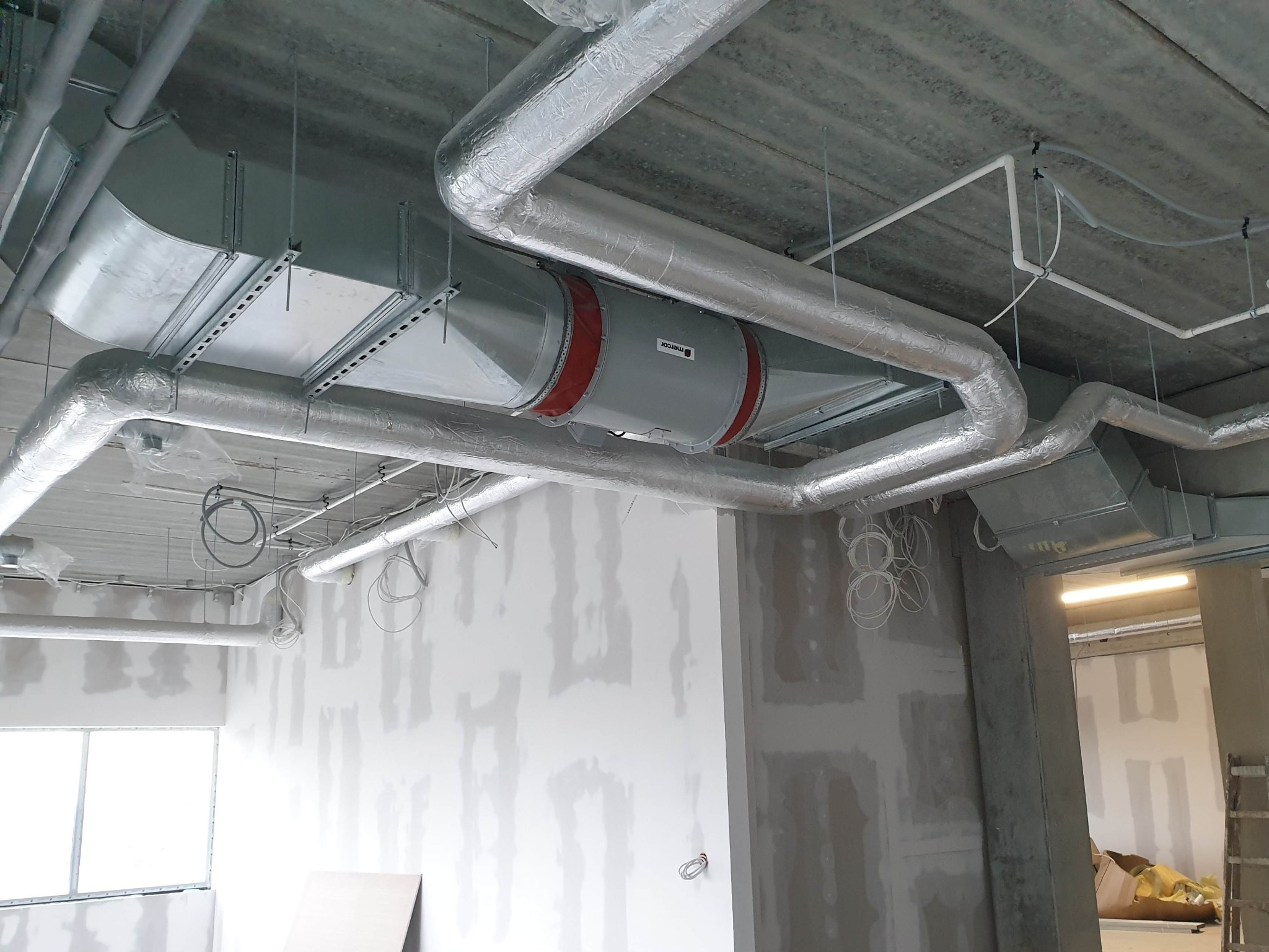 Montaż kanałów wentylacyjnych i rur izolowanych na suficie w surowym wnętrzu biurowym, widoczny element z logo Mercor.
