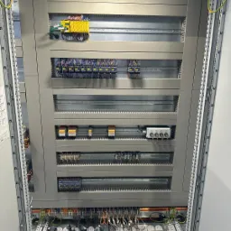 Otwarta szafa sterownicza z modułami elektrycznymi, widoczne liczne przewody, złącza, listwy montażowe i metalowa konstrukcja obudowy.