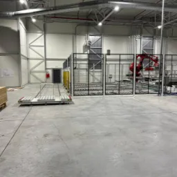 Wnętrze hali produkcyjnej z robotem przemysłowym za metalową siatką ochronną, widoczne rury instalacji pod sufitem i wózek transportowy na posadzce.