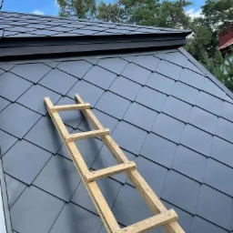 Nowy dach pokryty grafitową dachówką karpiówką, z drewnianą drabinką dekarską, widok z dołu na tle błękitnego nieba i zielonych drzew.