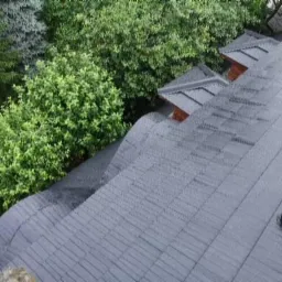 Widok z góry na ciemnoszary dach pokryty dachówką bitumiczną, z widocznymi kominkami wentylacyjnymi i lukarnami, otoczony zielenią drzew.