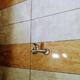 Bateria umywalkowa zamontowana na ścianie wyłożonej poziomymi pasami płytek w odcieniach beżu i kremu, z widocznymi fugami.