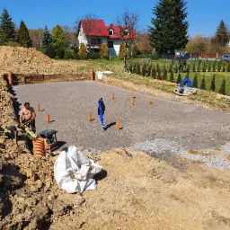 Przygotowanie podłoża pod fundamenty: ubijanie zagęszczarką wibro płytową, widoczne rury kanalizacyjne i geodezyjny niwelator laserowy w tle.