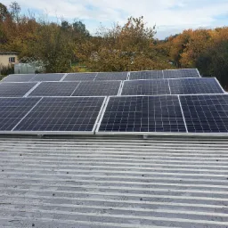 Instalacja fotowoltaiczna na falistym, metalowym dachu z widokiem na jesienne drzewa i budynek w tle.