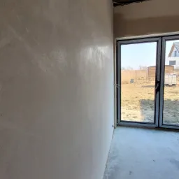 Świeżo nałożony, gładki tynk maszynowy na ścianie wewnętrznej, widoczny zarys okna balkonowego i surowy stan podłogi w pomieszczeniu.