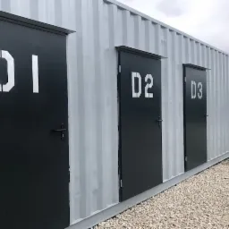 Zewnętrzny widok rzędu szarych kontenerów magazynowych z czarnymi drzwiami oznaczonymi białymi szablonowymi numerami D1, D2 i D3.
