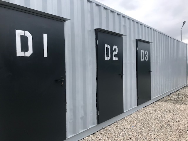 Zewnętrzny widok rzędu szarych kontenerów magazynowych z czarnymi drzwiami oznaczonymi białymi szablonowymi numerami D1, D2 i D3.