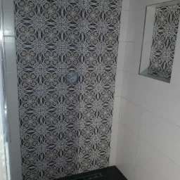 Wykończona kabina prysznicowa z dekoracyjnymi płytkami w odcieniach bieli, szarości i błękitu na ścianie oraz czarną, kwadratową kratką odpływową w podłodze.