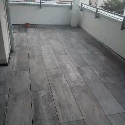 Balkon wyłożony szarymi, drewnopodobnymi płytkami ceramicznymi w podłużnym formacie, widoczny fragment ściany balkonu i metalowa barierka, w tle zabudowa jednorodzinna.